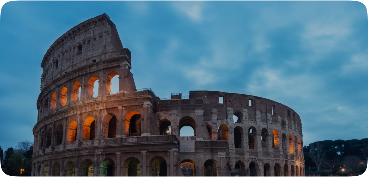 Imagem do Coliseu de Roma, uma estrutura circular com diversos arcos em suas entradas e janelas dispostas por toda a estrutura, feita de tijolos acinzentados teto aberto.